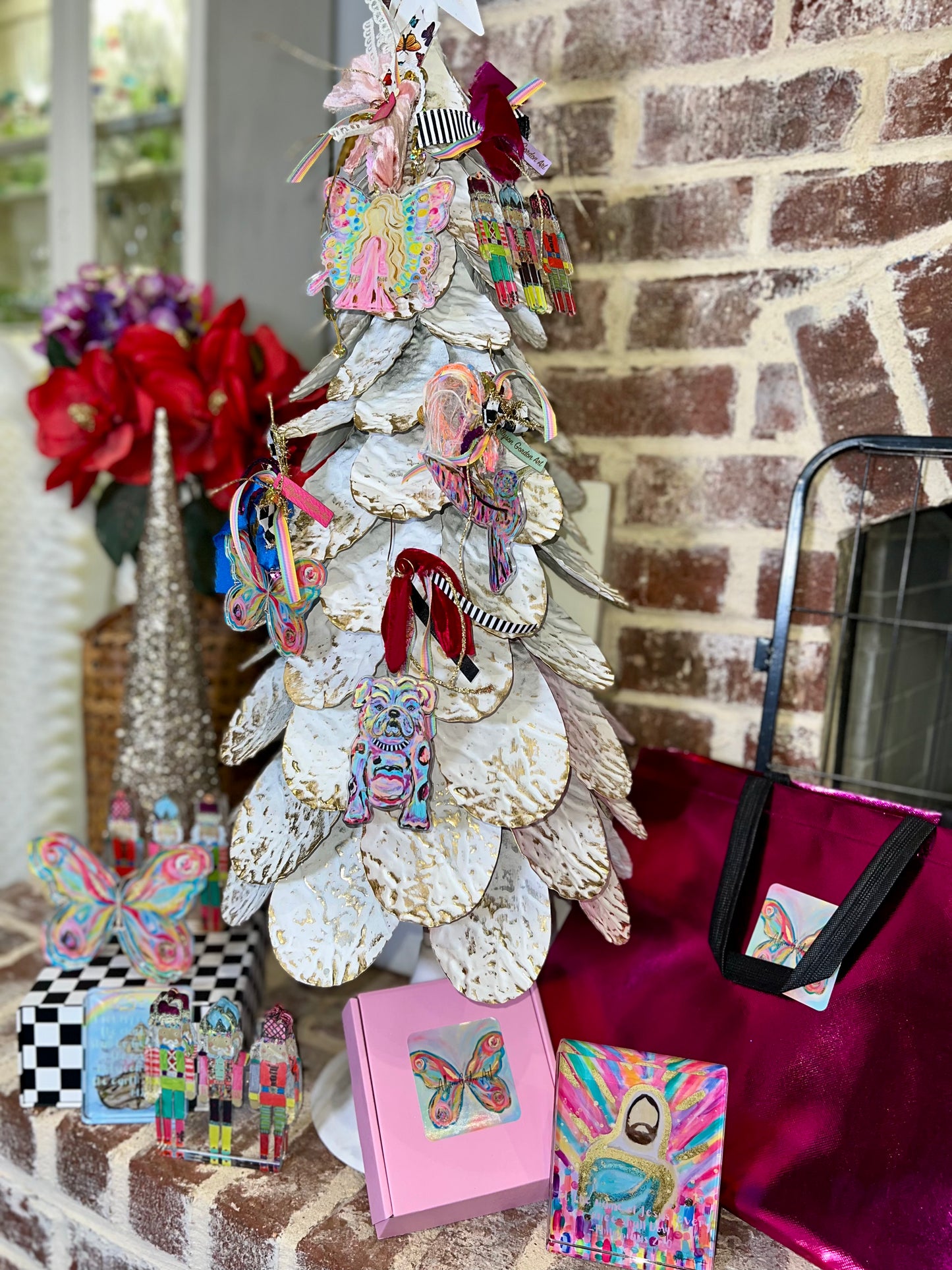 A decorative white Christmas tree with layered paper leaves and colorful handmade ornaments, styled on a brick surface with matching colorful gift boxes, a pink tote bag, and holiday flowers in the background.