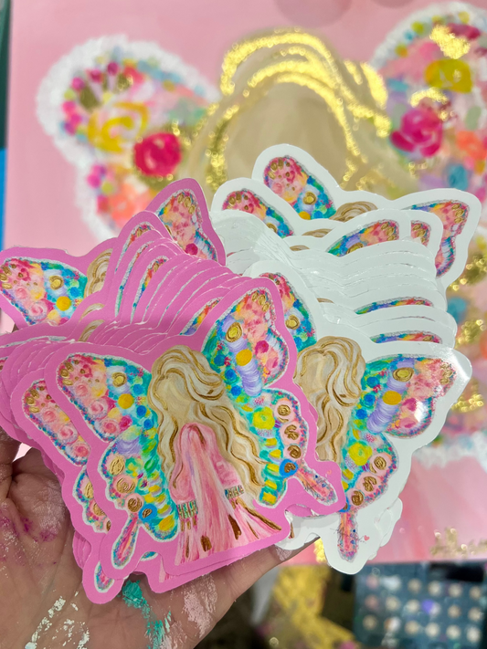 A hand holds a stack of colorful butterfly-shaped stickers featuring a fairy illustration with pink, blue, yellow, and green details. A pink box with matching butterfly artwork is visible in the background.